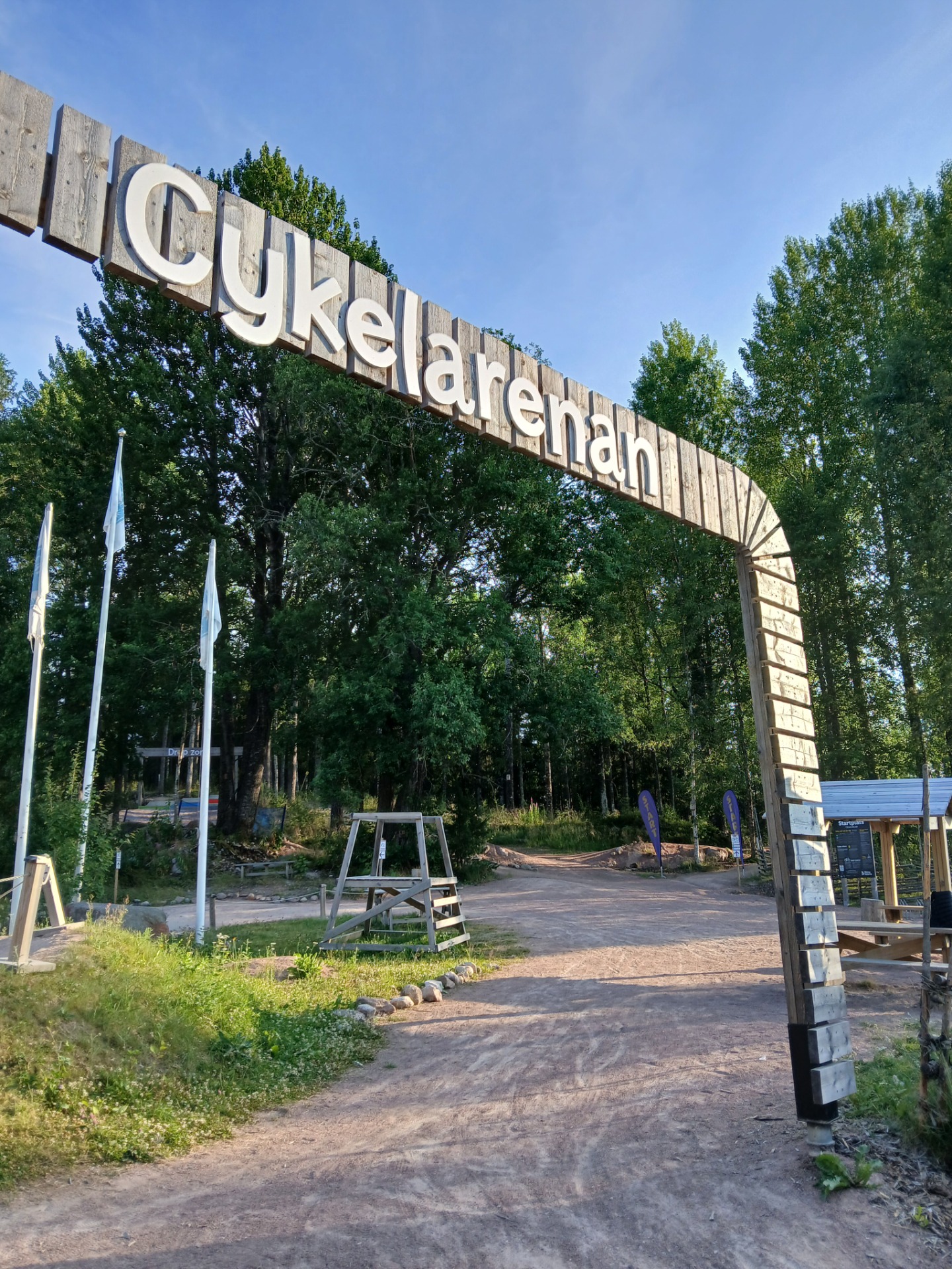 Stort træskilt med teksten Cykkelaren, omgivet af skov, grusvej og flag under en klar blå himmel.